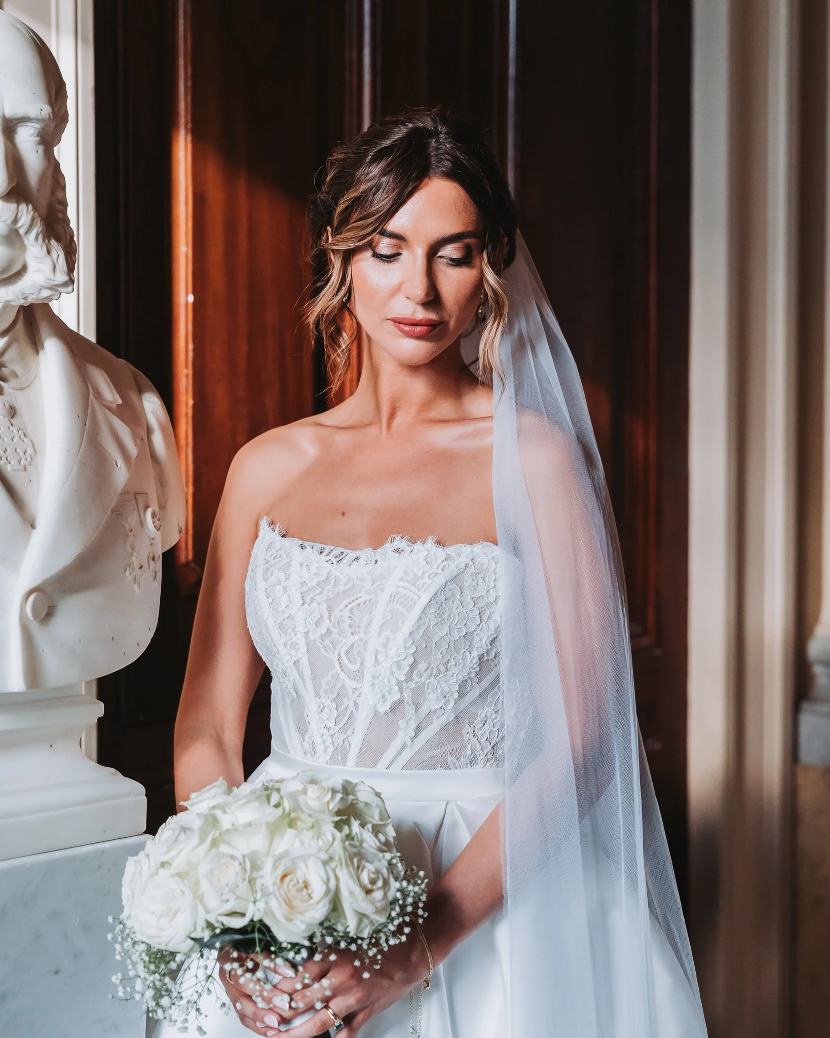A bride with shoulder-length brown hair styled in soft waves, wearing a white lace wedding dress and a veil, holding a bouquet of white flowers and standing next to a white bust sculpture of a bearded man.