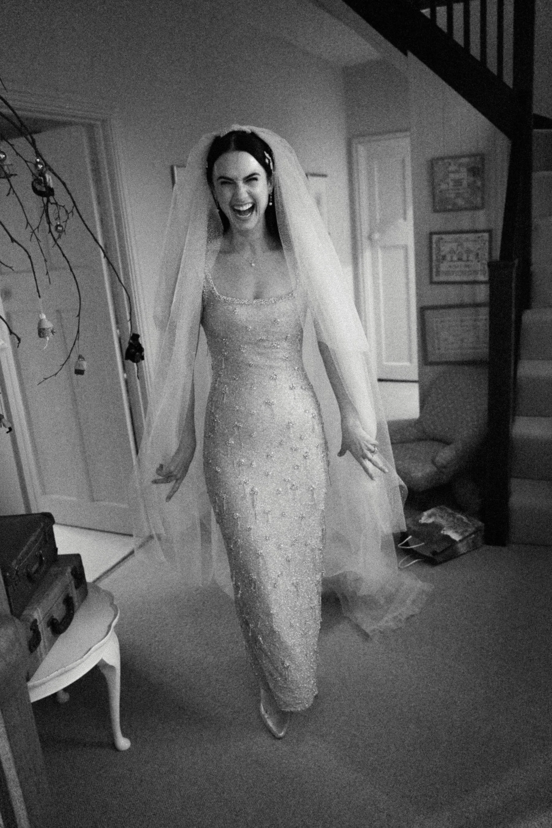 A woman dressed in a sparkly, form-fitting dress and veil, smiling and posing in a room with framed pictures on the wall and furniture around her.