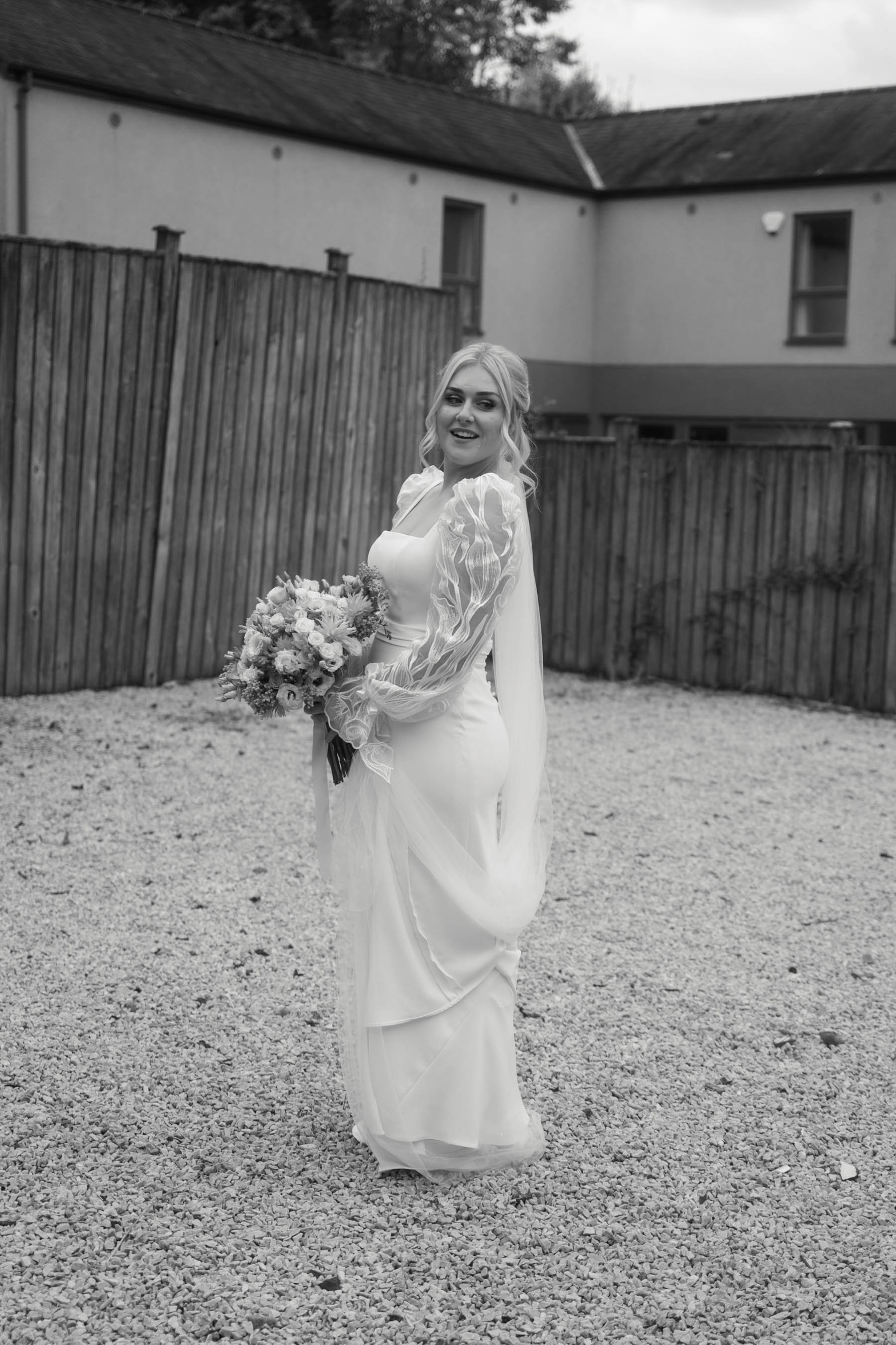 A smiling woman in a wedding dress holding a bouquet of flowers outdoors in front of a wooden fence.