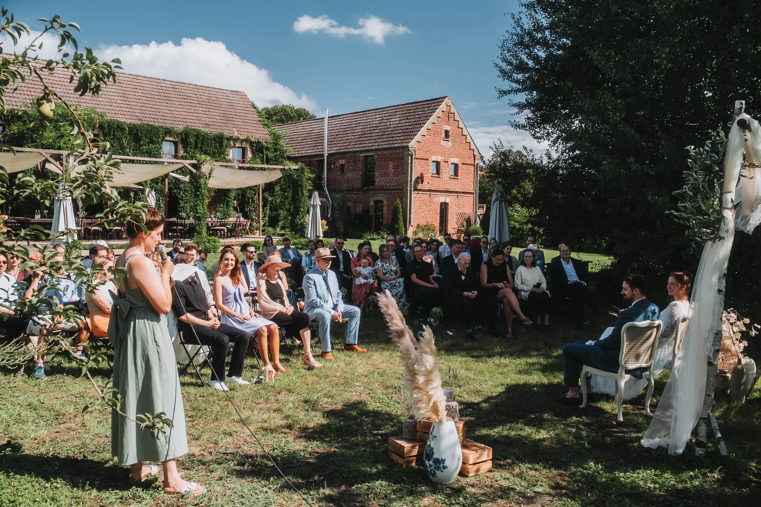 Outdoor wedding ceremony with guests seated on chairs, a woman reading at the front, and a decorated arch area in a garden setting with trees and brick buildings in the background.