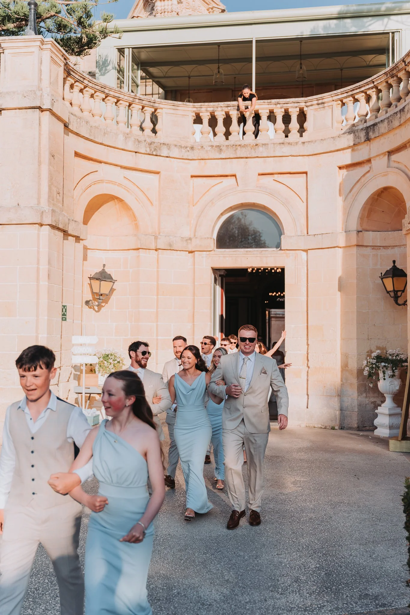 People dressed in formal attire, including a woman in a light blue dress and a man in a beige suit, walking outside a historic building with arches and stone walls, during a sunny day.