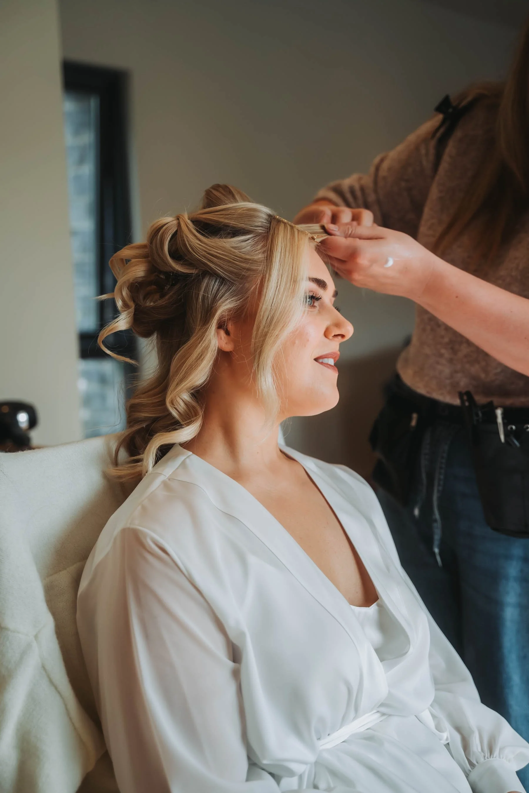 A woman with blond hair styled in loose curls, wearing a white robe, sits on a beige chair with a slight smile as a hair stylist works on her hair in a salon or studio.