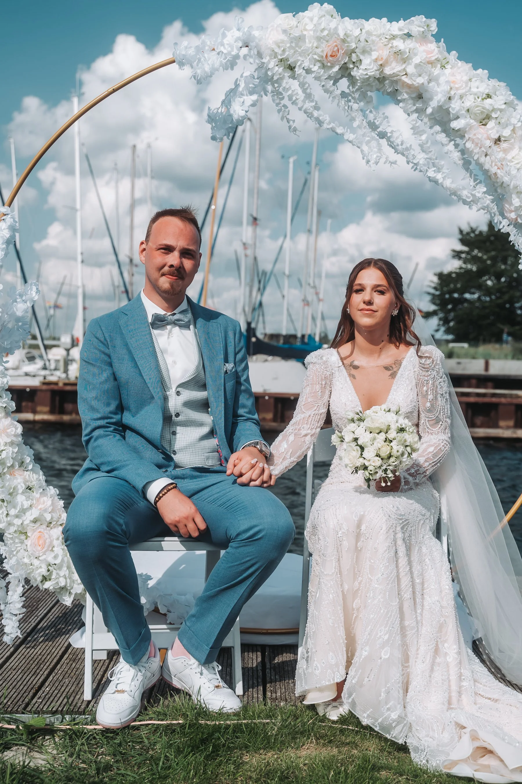 A bride and groom sitting together on a boat dock, holding hands, during their wedding celebration outdoors near a marina, with boats and clouds in the background.