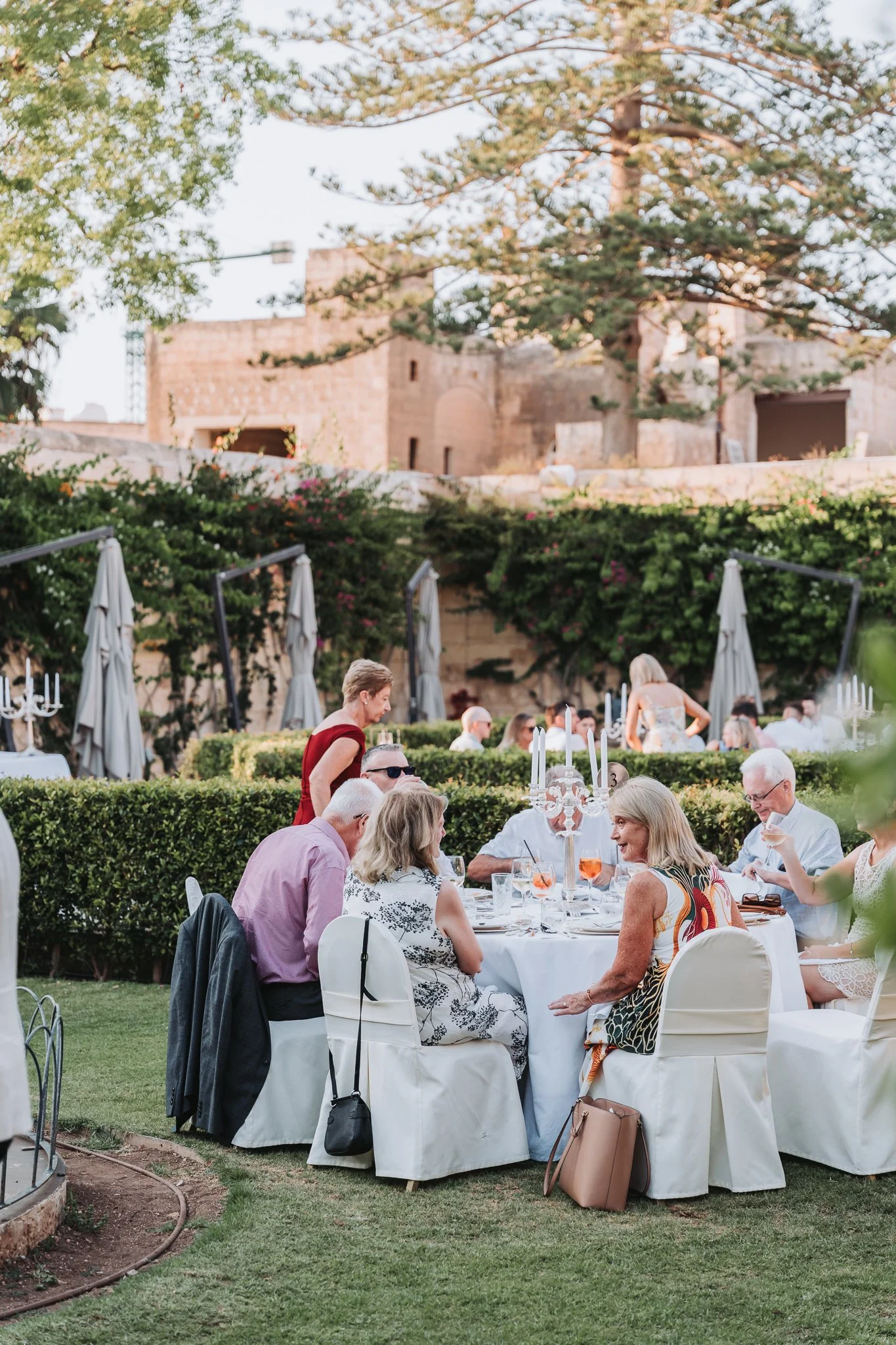 People dining outdoors at a formal event in a garden with a historic stone building and trees in the background.