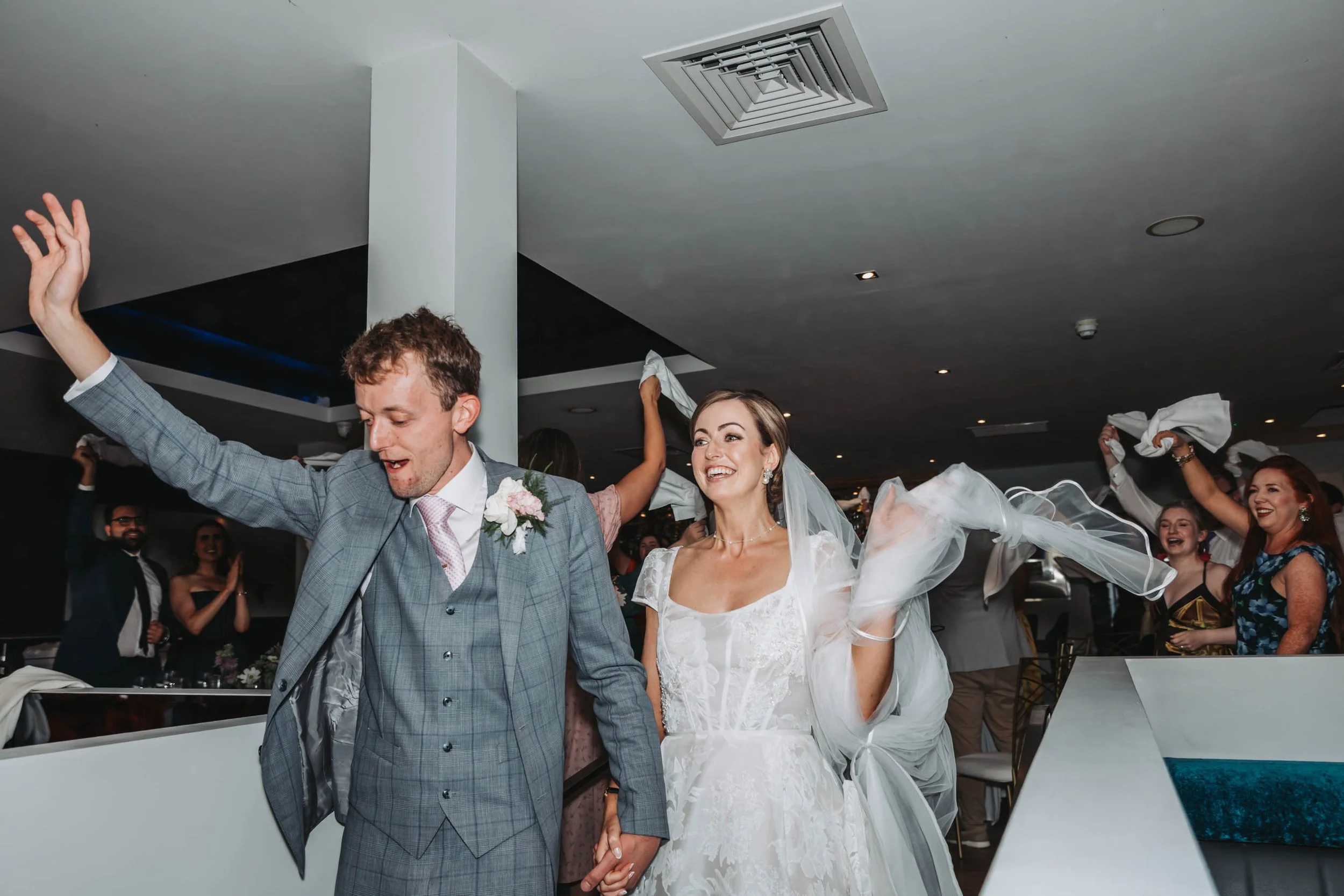 A wedding celebration with a bride in a white gown and a groom in a gray suit holding hands and dancing with guests in the background.