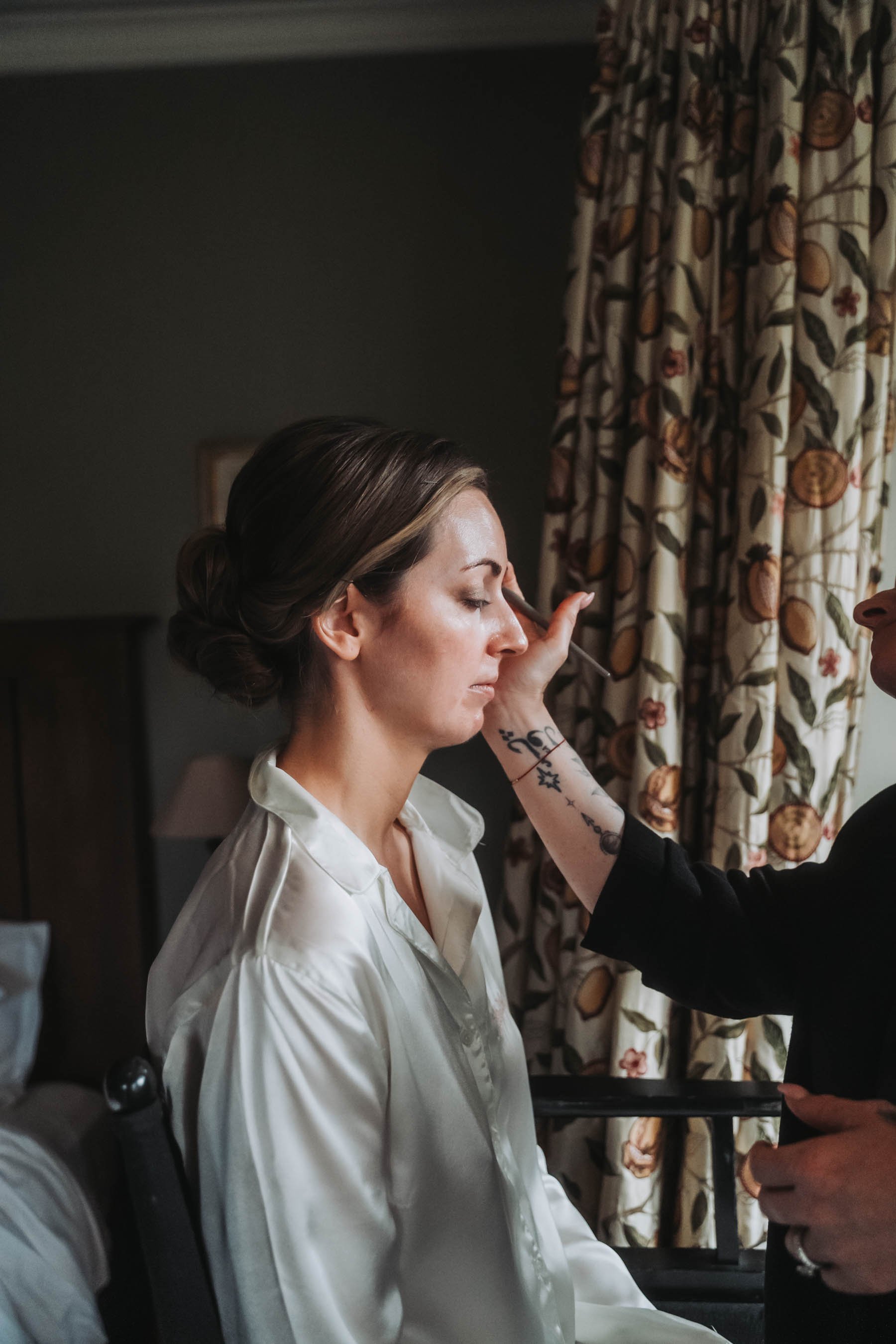 A woman with brown hair styled in a bun, wearing a white satin robe, is having makeup applied to her eyelid by a makeup artist with tattoos on their arm, in a room with patterned curtains.