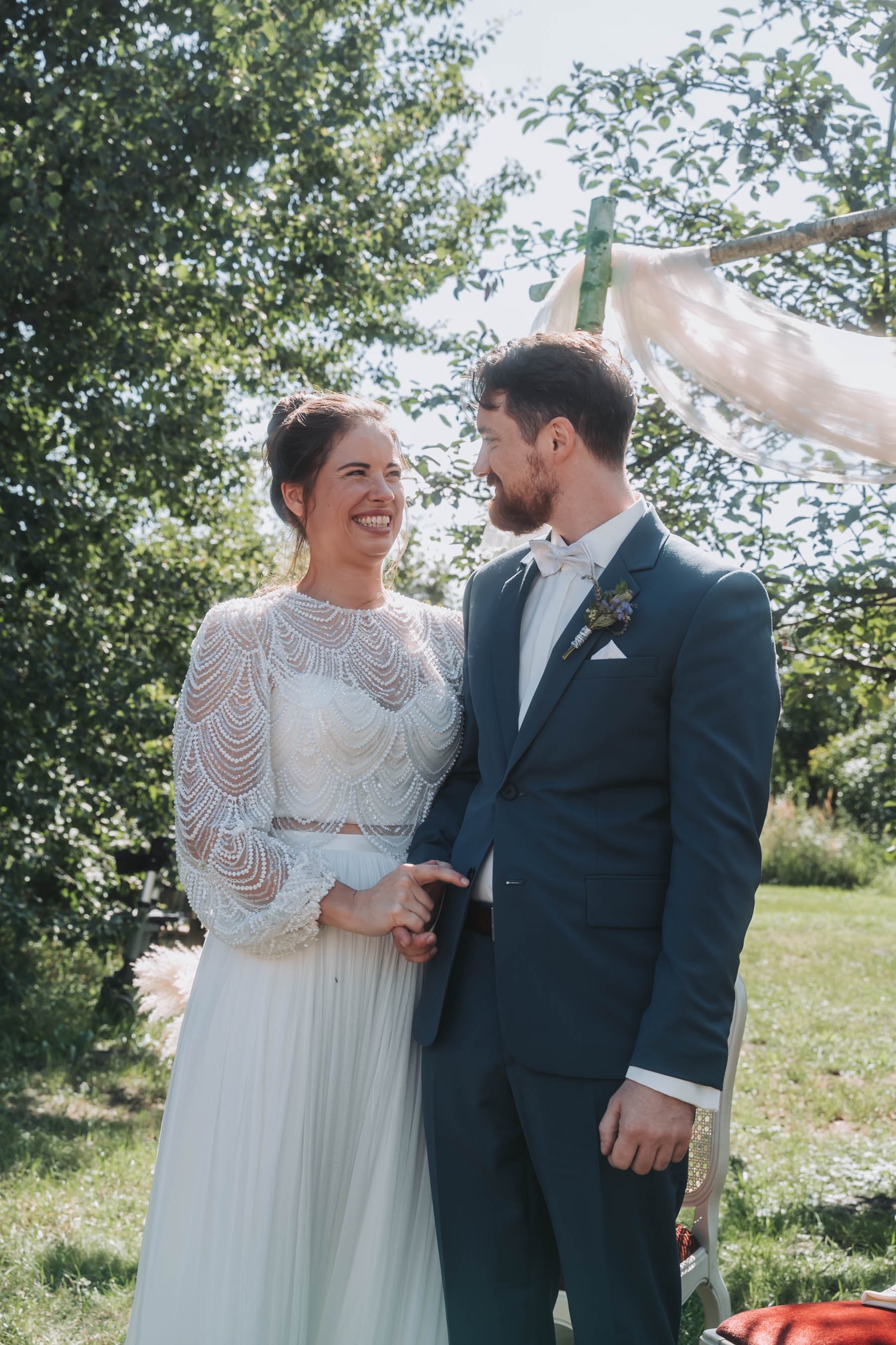 A bride and groom standing outdoors, holding hands, and looking at each other with happy expressions. The bride is wearing a white lace wedding dress with long sleeves, and the groom is dressed in a dark suit with a bow tie. The background features t