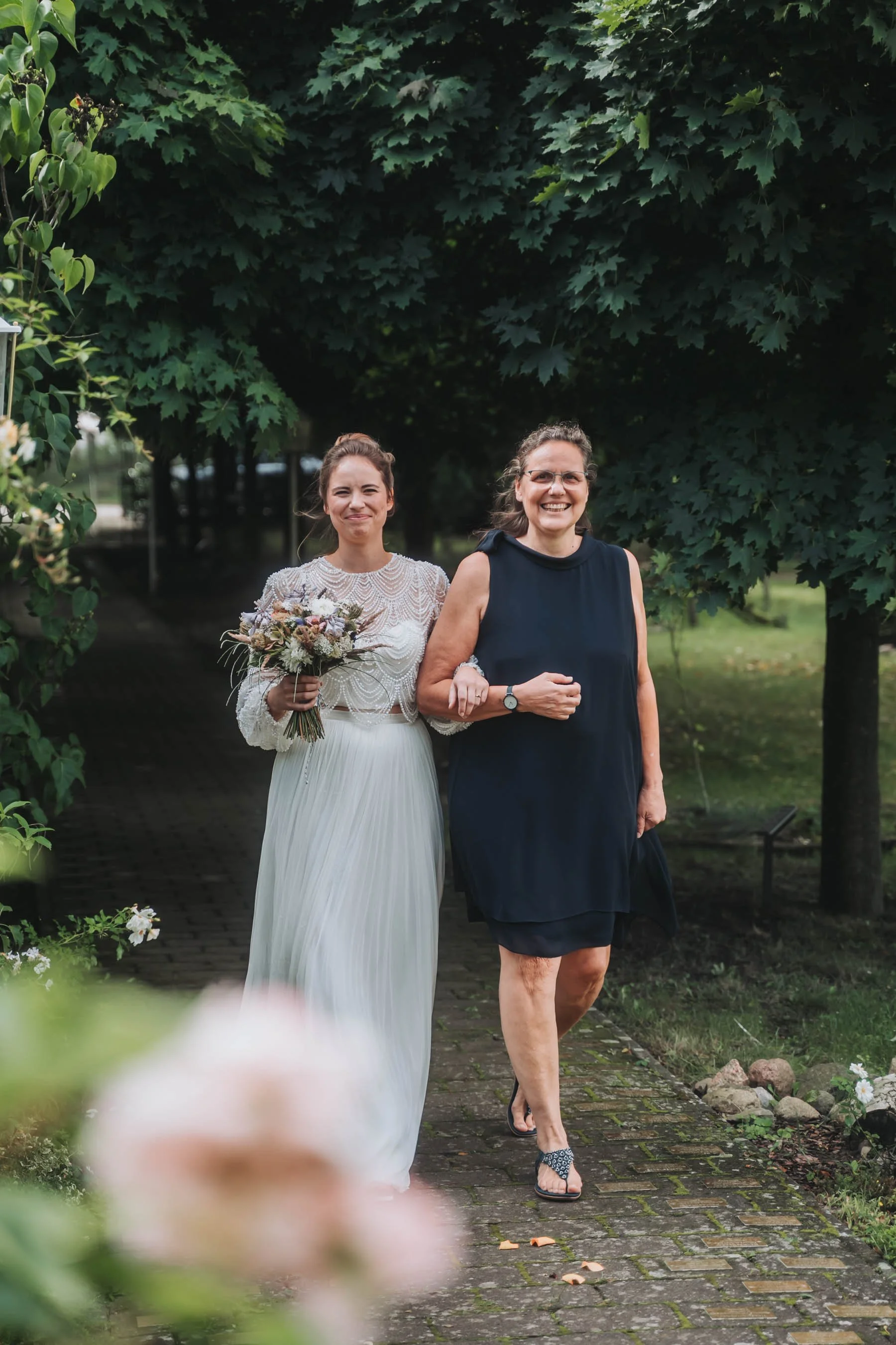 A bride in a white wedding dress with lace details walking down an outdoor garden path, holding a bouquet of flowers, accompanied by an older woman in a dark sleeveless dress, smiling.