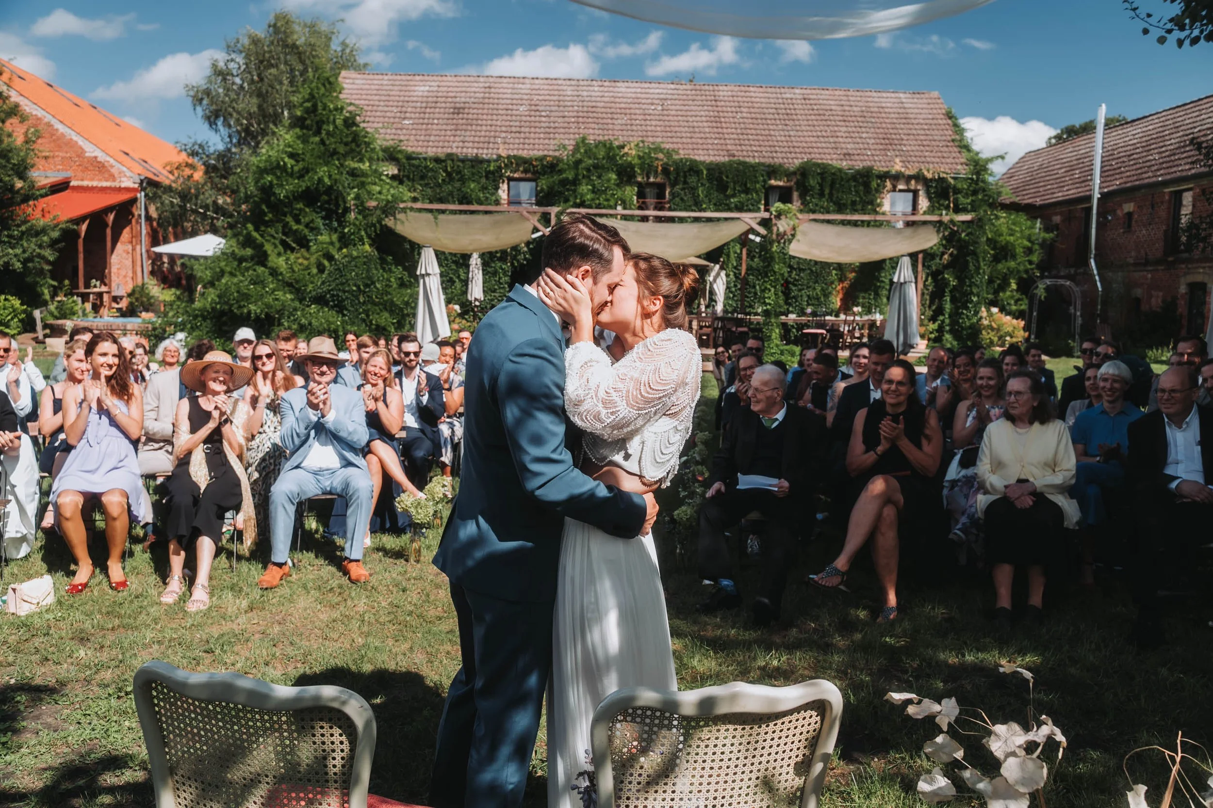 A couple holding each other and kissing during an outdoor wedding ceremony with guests watching, in a garden setting with a rustic house in the background.