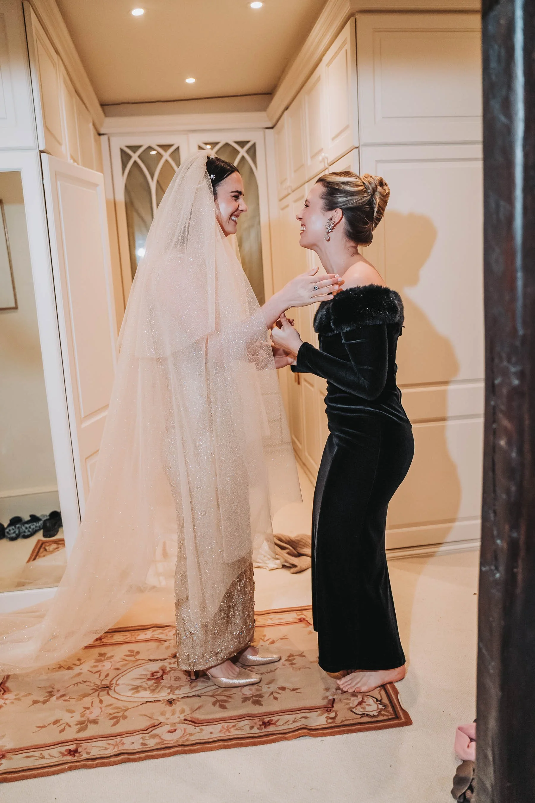 Two women are laughing and sharing a joyful moment in a room with beige cabinets. One is wearing a sparkly wedding dress and veil, while the other is dressed in a black gown with fur details and statement earrings.