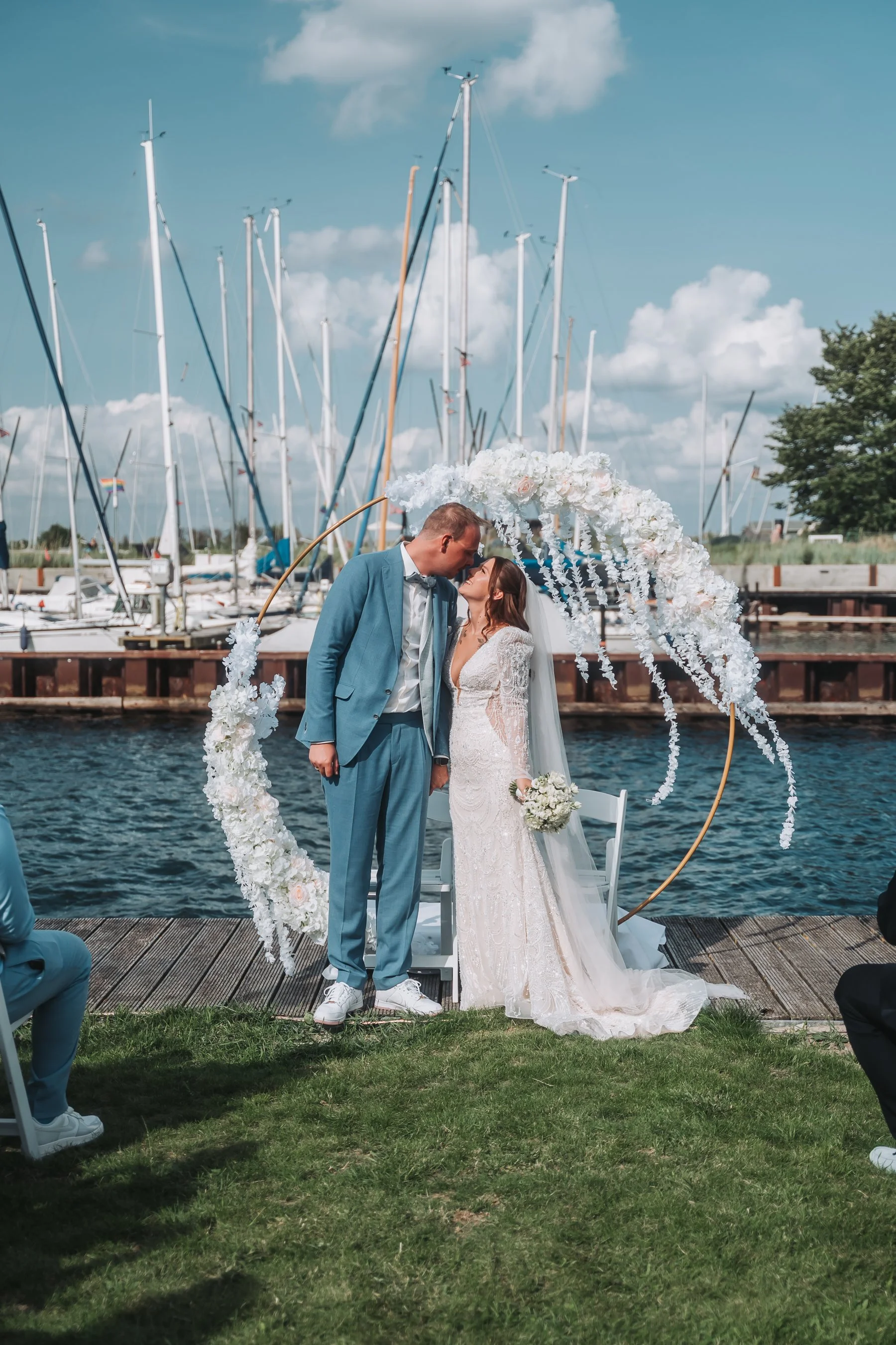 A bride and groom standing under a floral hoop arch near a body of water, with sailboats and marina in the background, during an outdoor wedding ceremony.