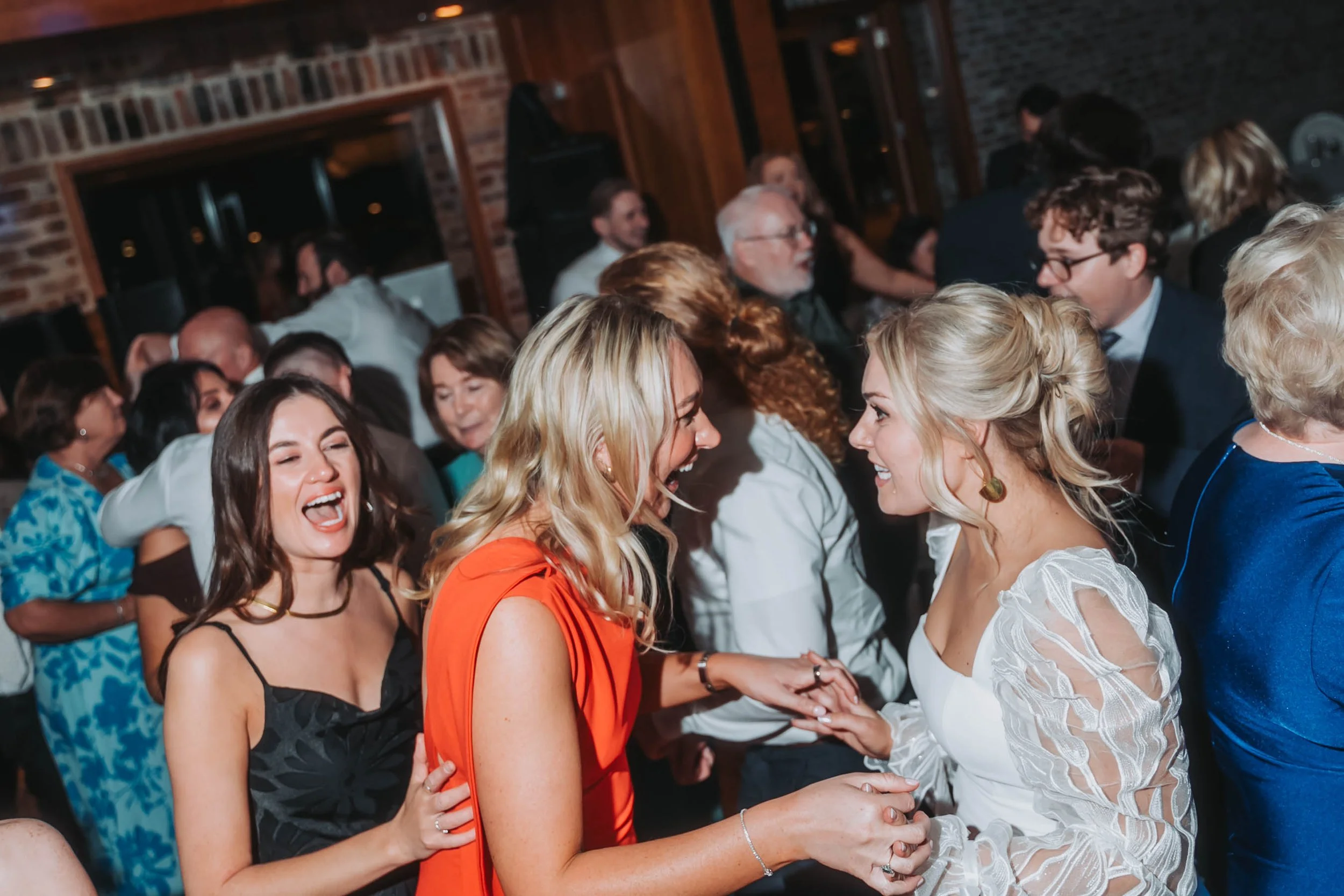 People dancing and smiling at a lively party or celebration in a cozy, brick-walled venue.