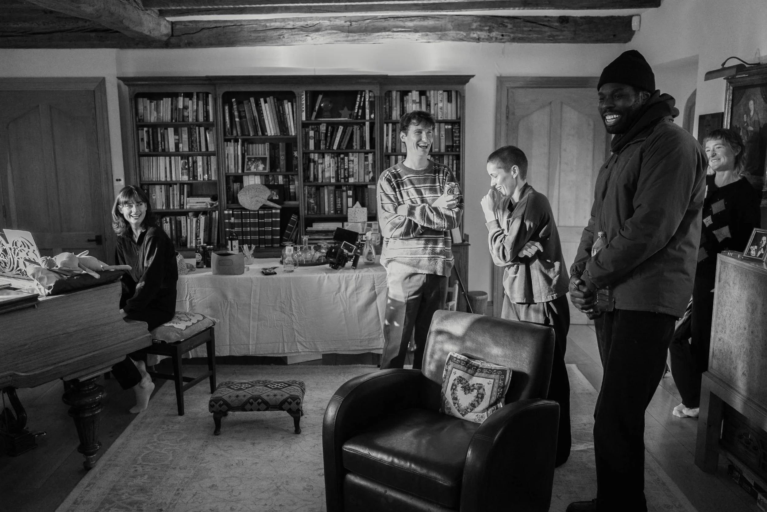 People gathered in a cozy room, smiling and engaging in conversation. There are bookshelves filled with books, a table with various items, and a mix of chairs and a piano, indicating a warm, social atmosphere.