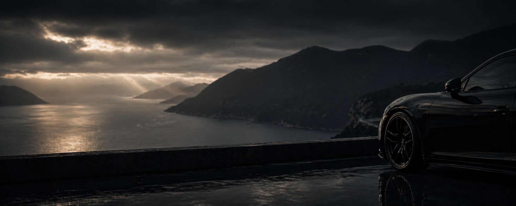 A black sports car parked on a mountain road overlooking a body of water with mountains in the background during a cloudy, overcast day.
