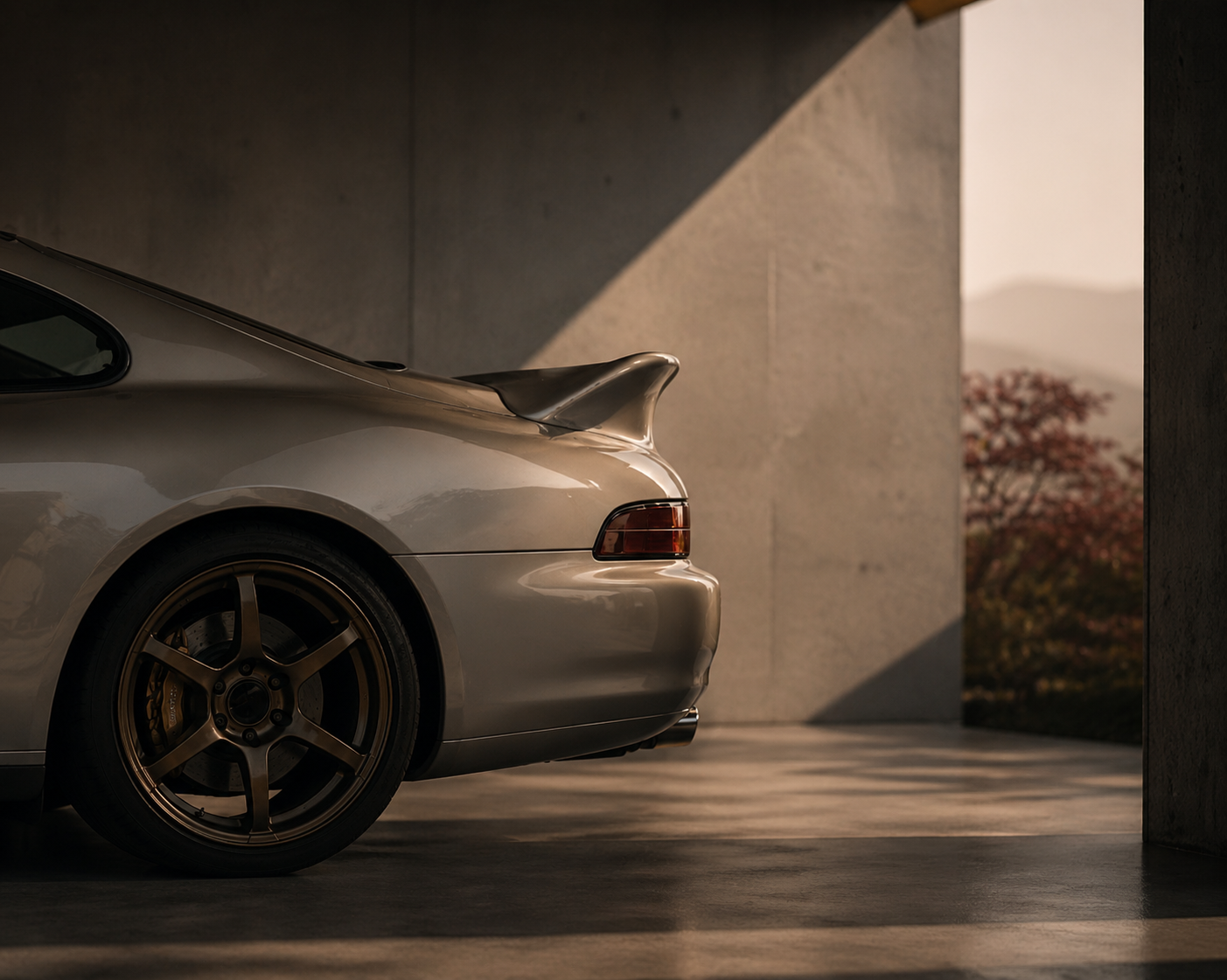 Partial view of a silver sports car parked indoors with a dark background, focusing on the rear quarter panel, wheel, and spoiler, with a glimpse of an outdoor scene with trees in the distance.
