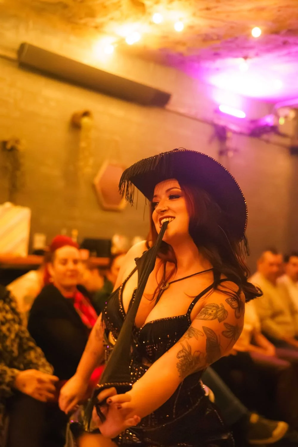 A woman dressed in black with tattoos on her arm, wearing a large black cowboy hat, is pulling off a glove with her teeth at a lively indoor gathering with pink and yellow lighting.
