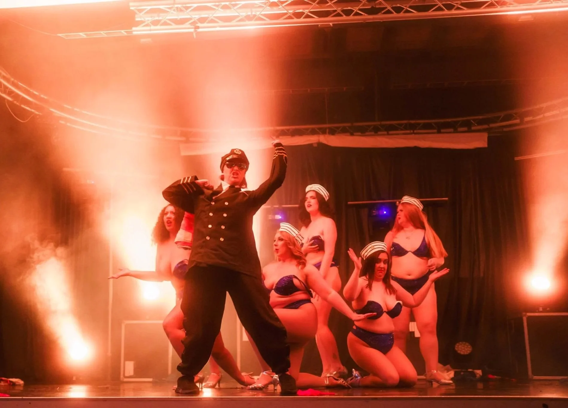 Performer in a black uniform dancing on stage with women dressed in blue bikinis, some kneeling and others standing, under warm stage lighting.