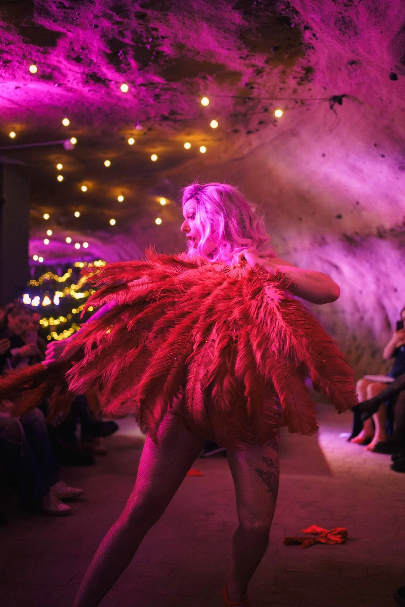 A performer on stage wearing a costume with large red and pink feathers, pink wigs, fishnet tights, and high heels. The stage is illuminated with purple and yellow hanging lights, and background market or festival decorations.