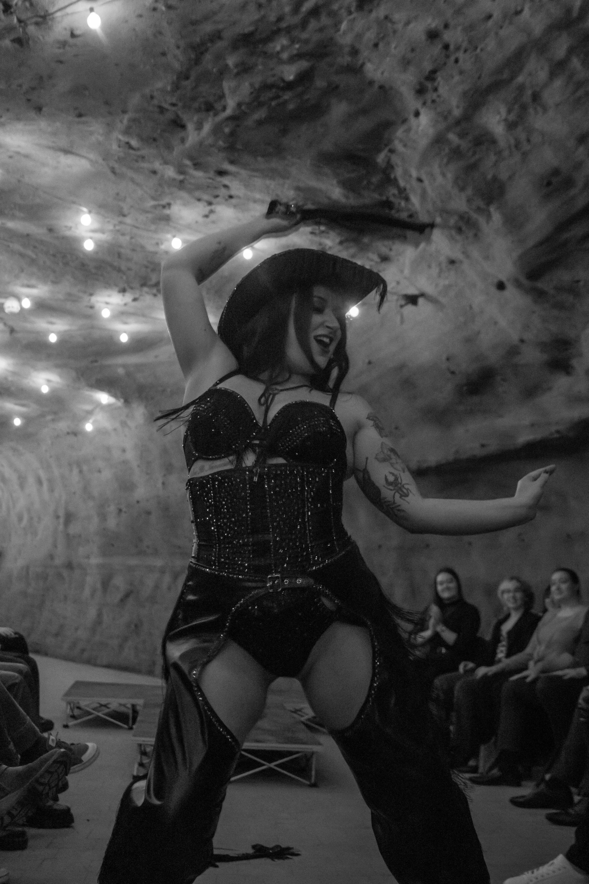 A woman in a black outfit and wide-brimmed hat performs a dance move, smiling, inside a cave-like venue with string lights and an audience watching.