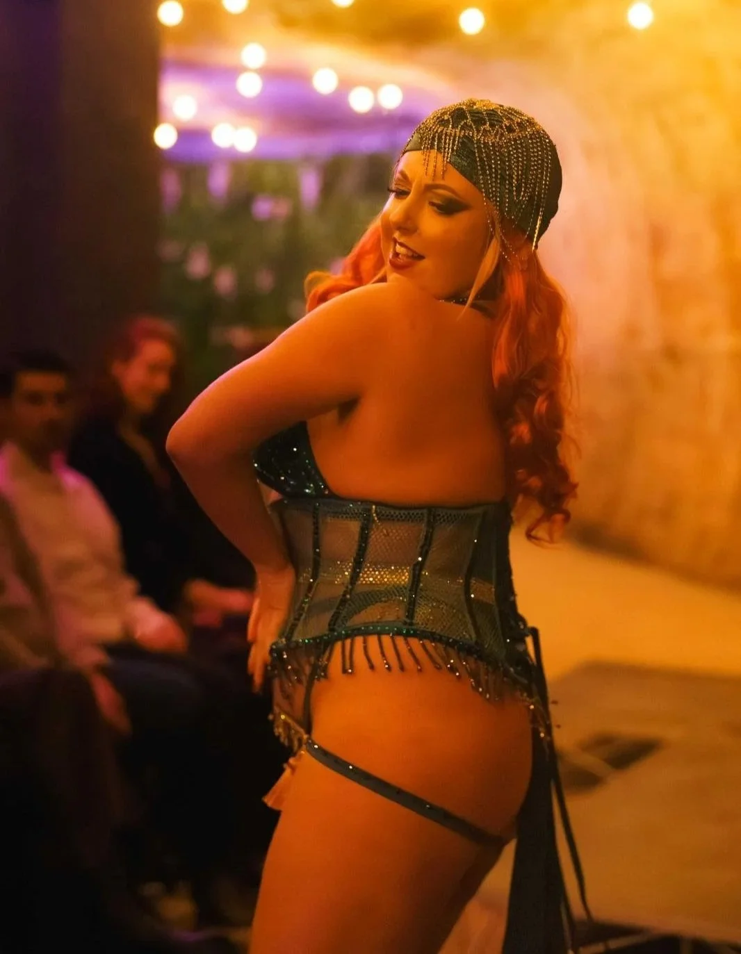 A performer in a black and sparkly costume with a beaded headpiece dances on stage in a dimly lit environment with colorful lights.