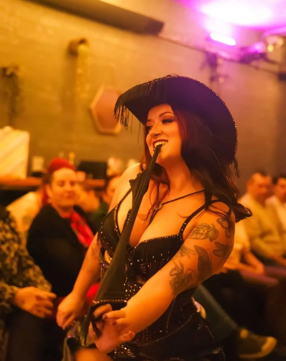 A woman wearing a black cowboy hat biting an umbrella at a lively indoor event, with a background of seated people and purple neon lighting.