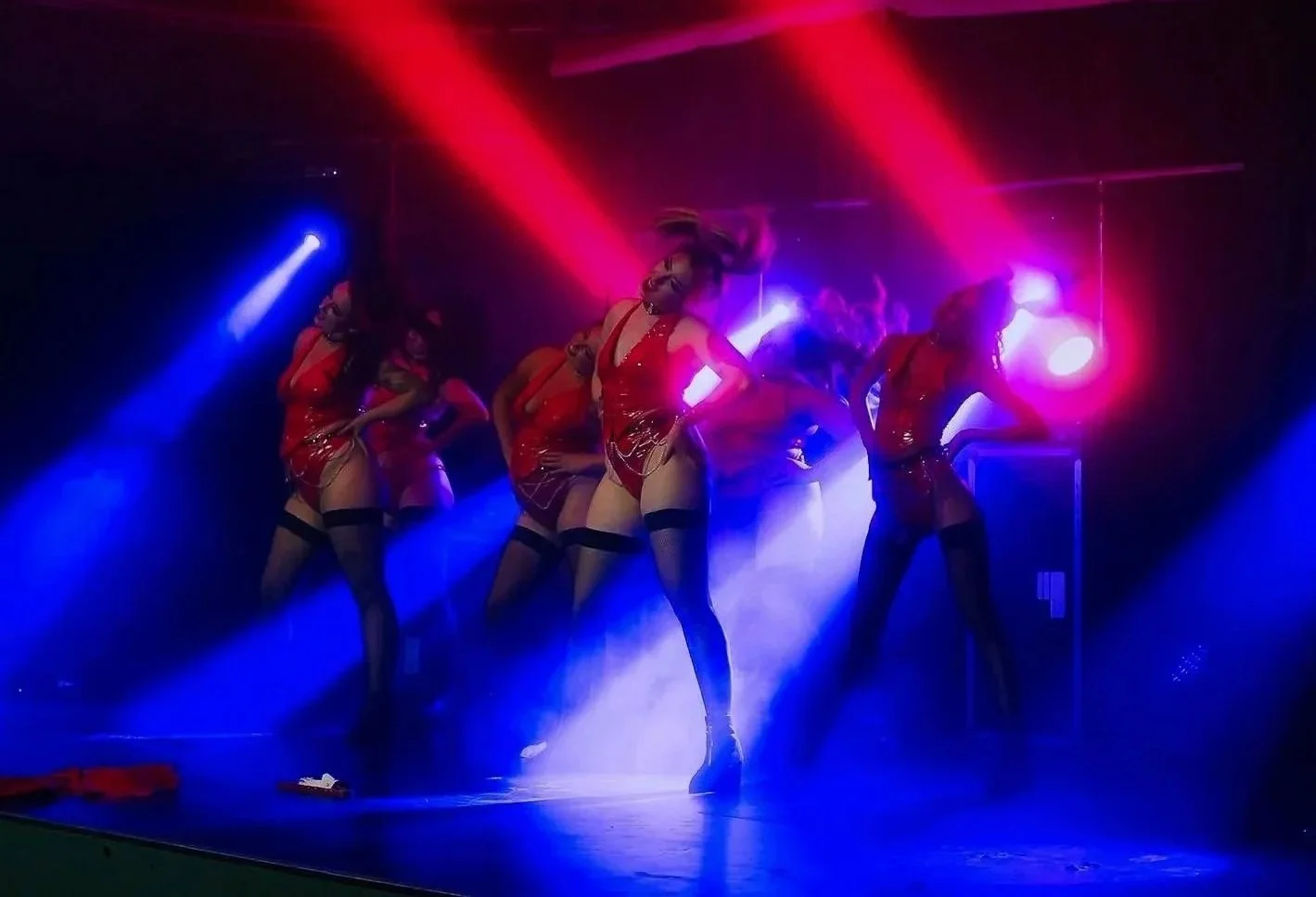 A group of female dancers performing a stage dance routine in red costumes, with colorful stage lighting and smoke effects.