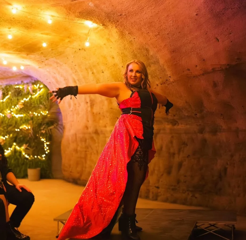 Woman in a red and black dress dancing in a warmly lit tunnel with string lights and a decorated Christmas tree in the background.