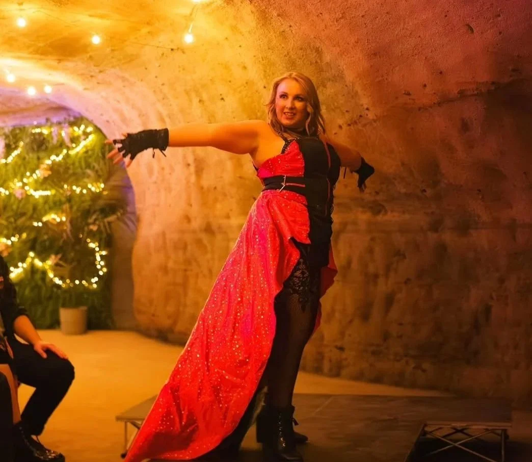 A woman in a red and black dress with lace details, posing with her arms outstretched inside a tunnel with brick walls and a decorated Christmas tree in the background.