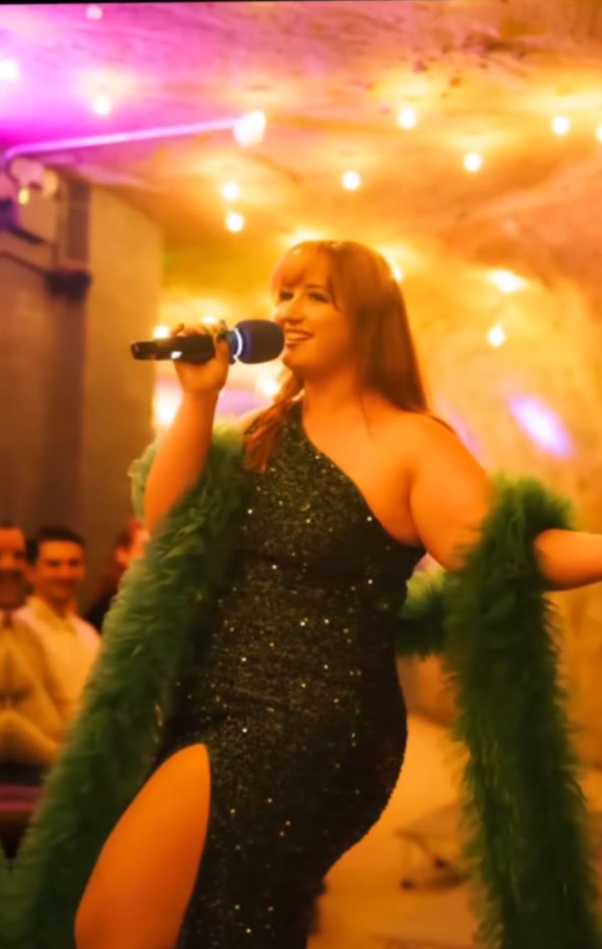 Woman singing into a microphone at a party, wearing a sparkly black dress and a green feather boa, with colorful string lights and guests in the background.