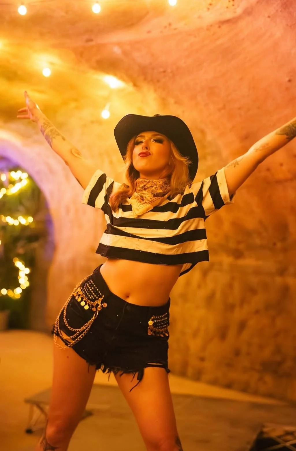 A woman with light skin, wavy hair, and tattoos, wearing a black cowboy hat, striped crop top, and black shorts with chains, standing with arms outstretched in a warm, dimly lit setting with a brick arch ceiling and fairy lights in the background.