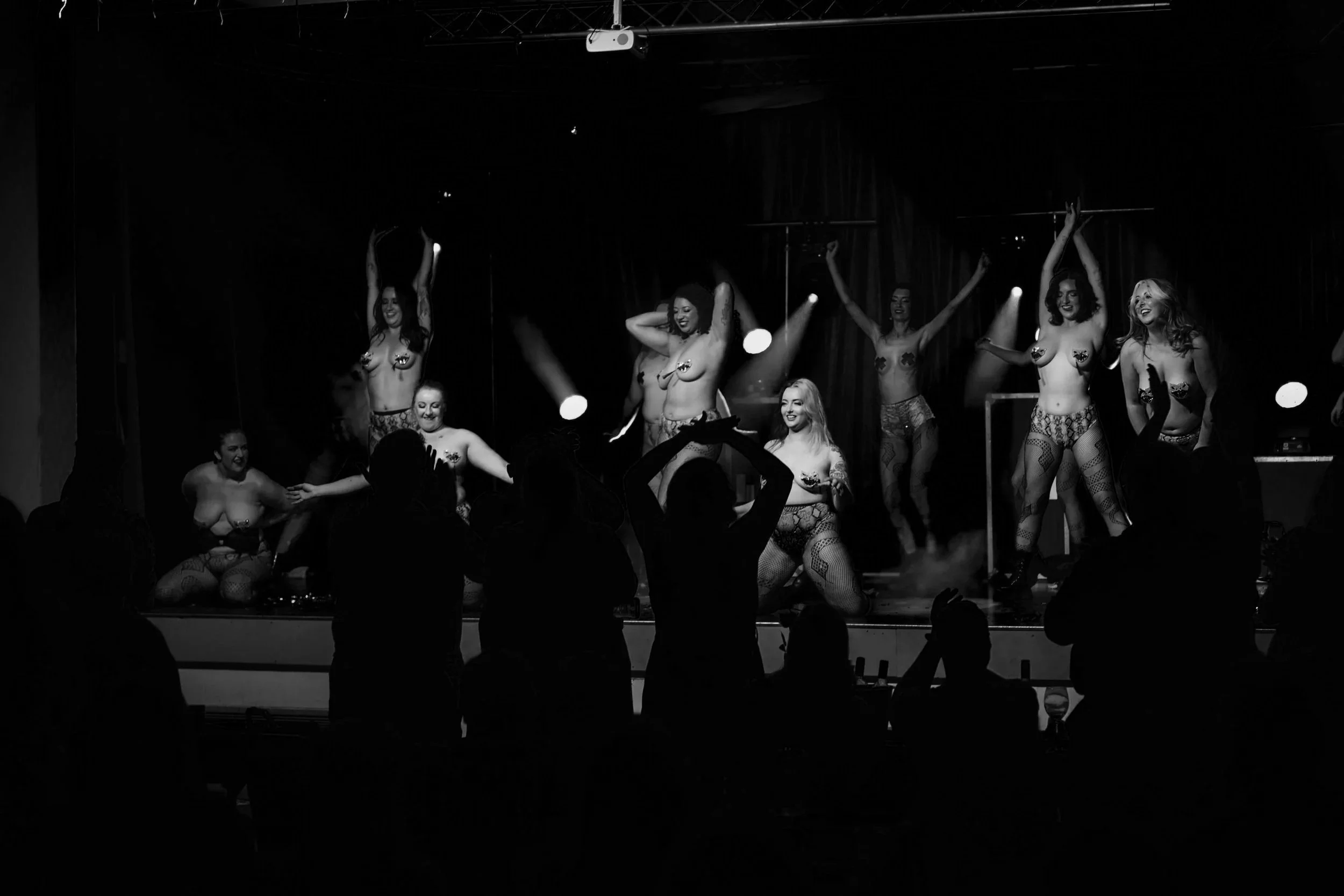 A group of performers on stage, dressed in lingerie, with some holding feather fans and others kneeling or standing, under stage lights.