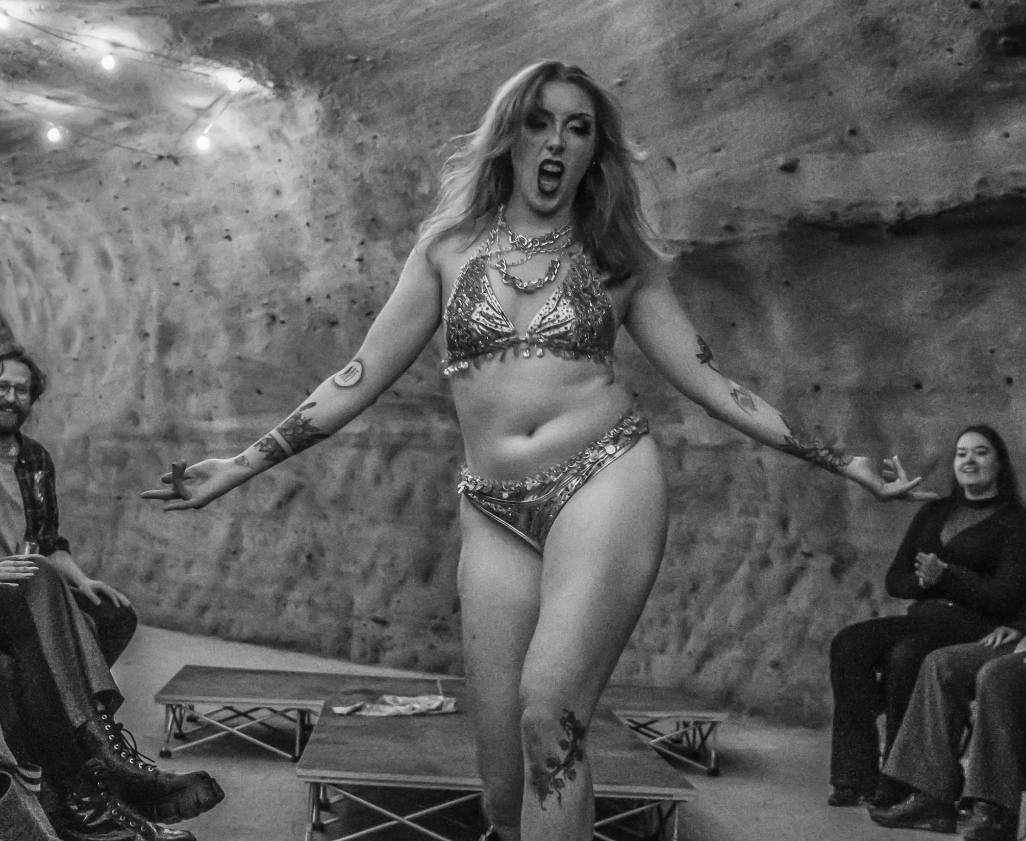 A woman with tattoos and curly hair, wearing a shiny metallic bikini top and jewelry, is standing with her mouth open in a dramatic pose in front of a rock or cave wall. People are seated around her, some smiling and watching her perform.