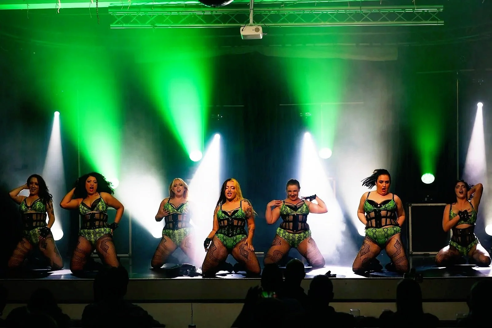 A group of seven female performers on stage dressed in green and black costumes, dancing with colorful stage lighting including white and green beams. Audience silhouettes are visible in the foreground.