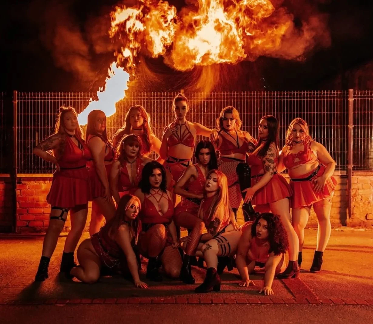 A group of women dressed in red outfits, posing in front of a fence with fire and flames in the background at night.