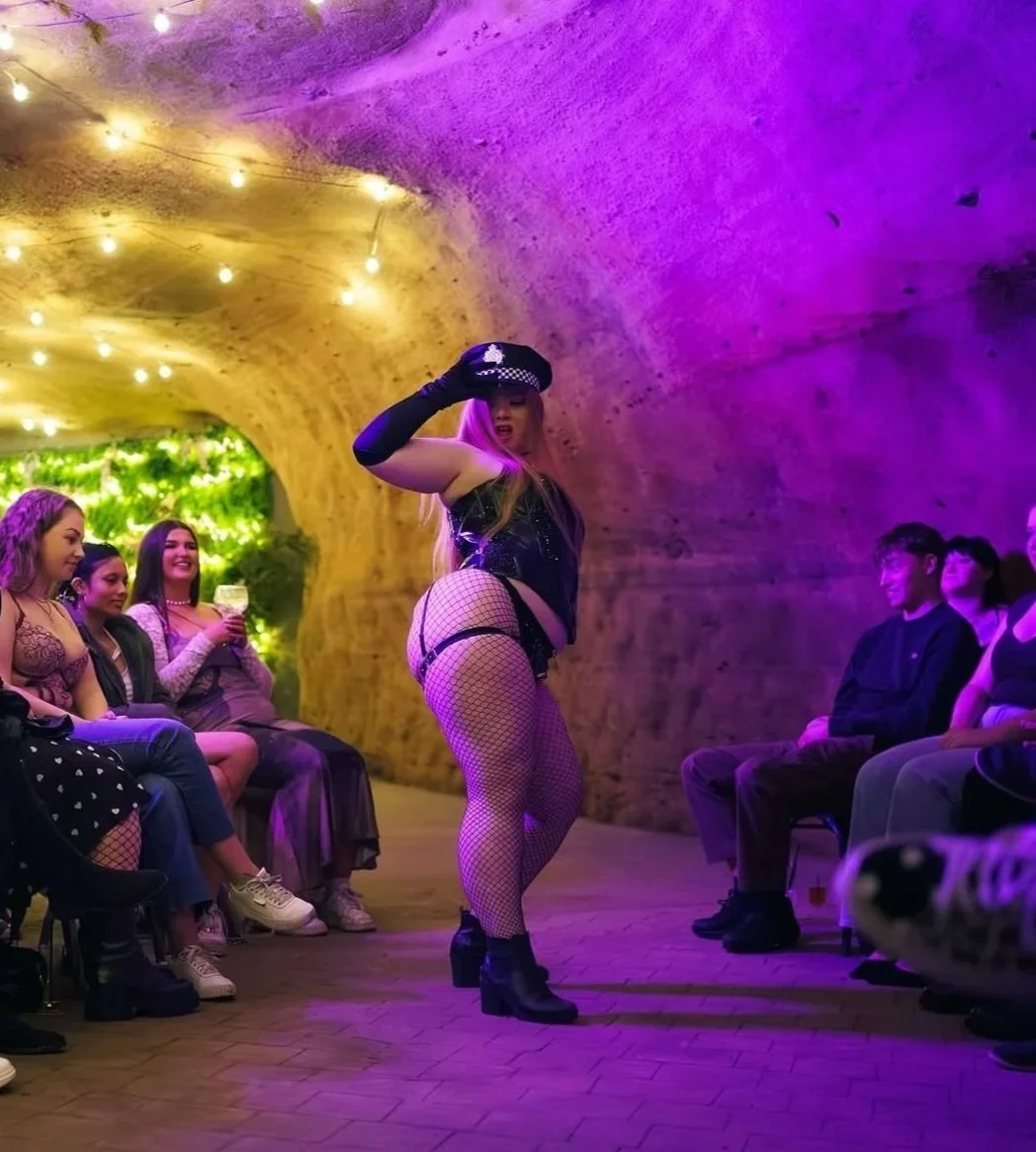 A performer dressed in black with fishnet stockings and police costume elements dancing in a tunnel with purple and yellow lighting, while an audience of women and men watch and smile.