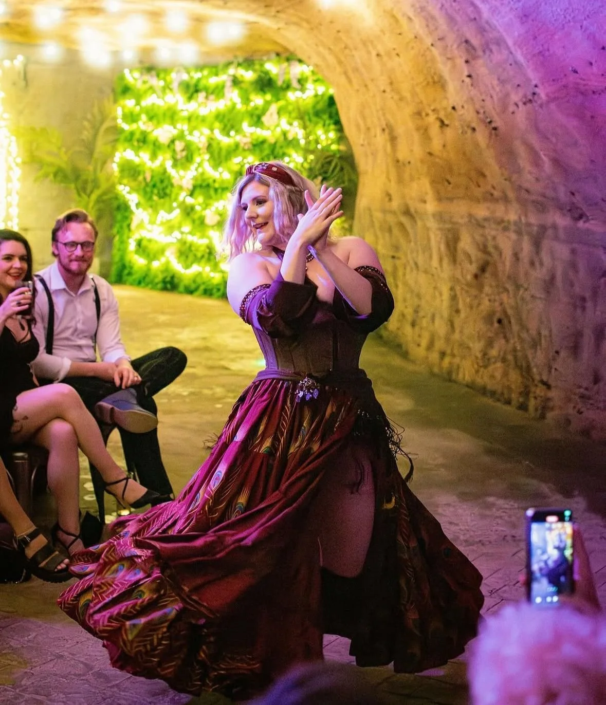 A woman in a colorful, flowing dress dances in a cave-like setting with green and purple lighting. Two people sit and watch her, and a person in the foreground takes a photo of the dancer.