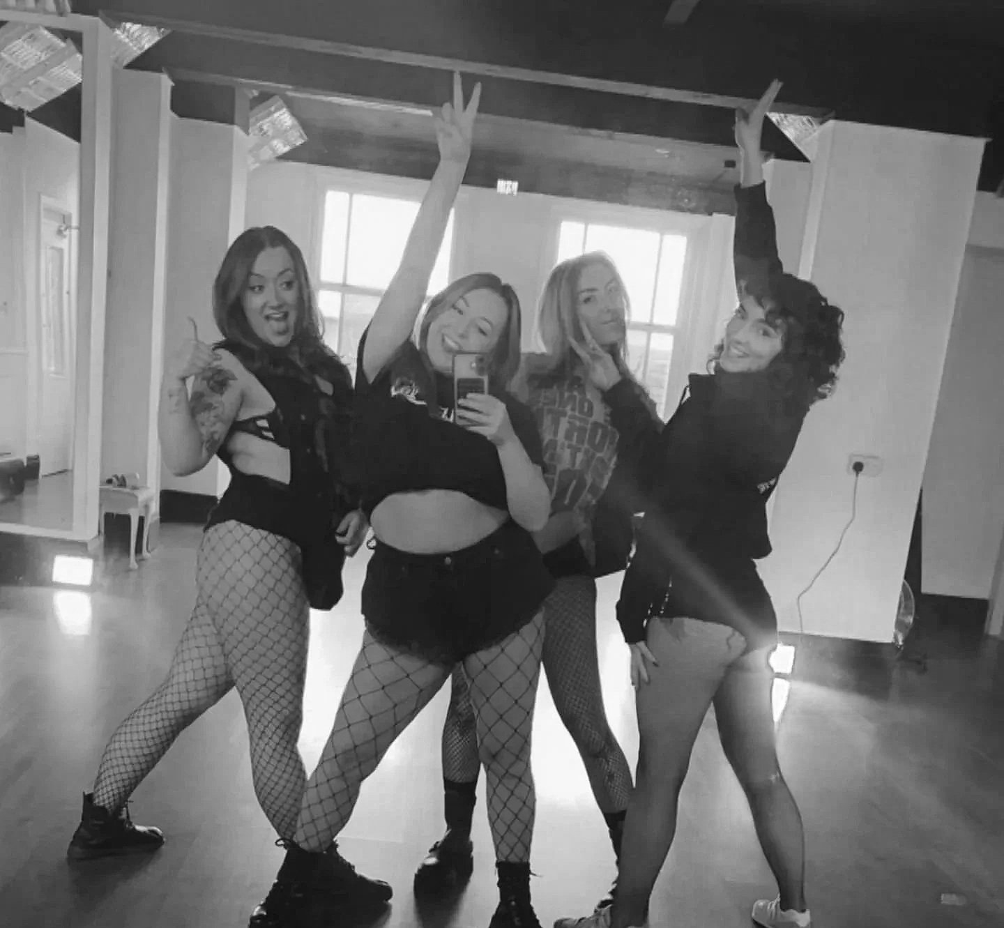 Four women posing for a mirror selfie in a room with large windows. They are dressed in edgy, revealing outfits with fishnet stockings and alternative fashion styles.