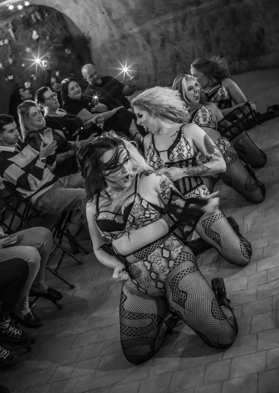 Two women in revealing, patterned outfits and fishnet stockings are dancing on the floor at a party or nightclub, with an audience of mostly women watching, some clapping and smiling. The scene is lively and energetic.