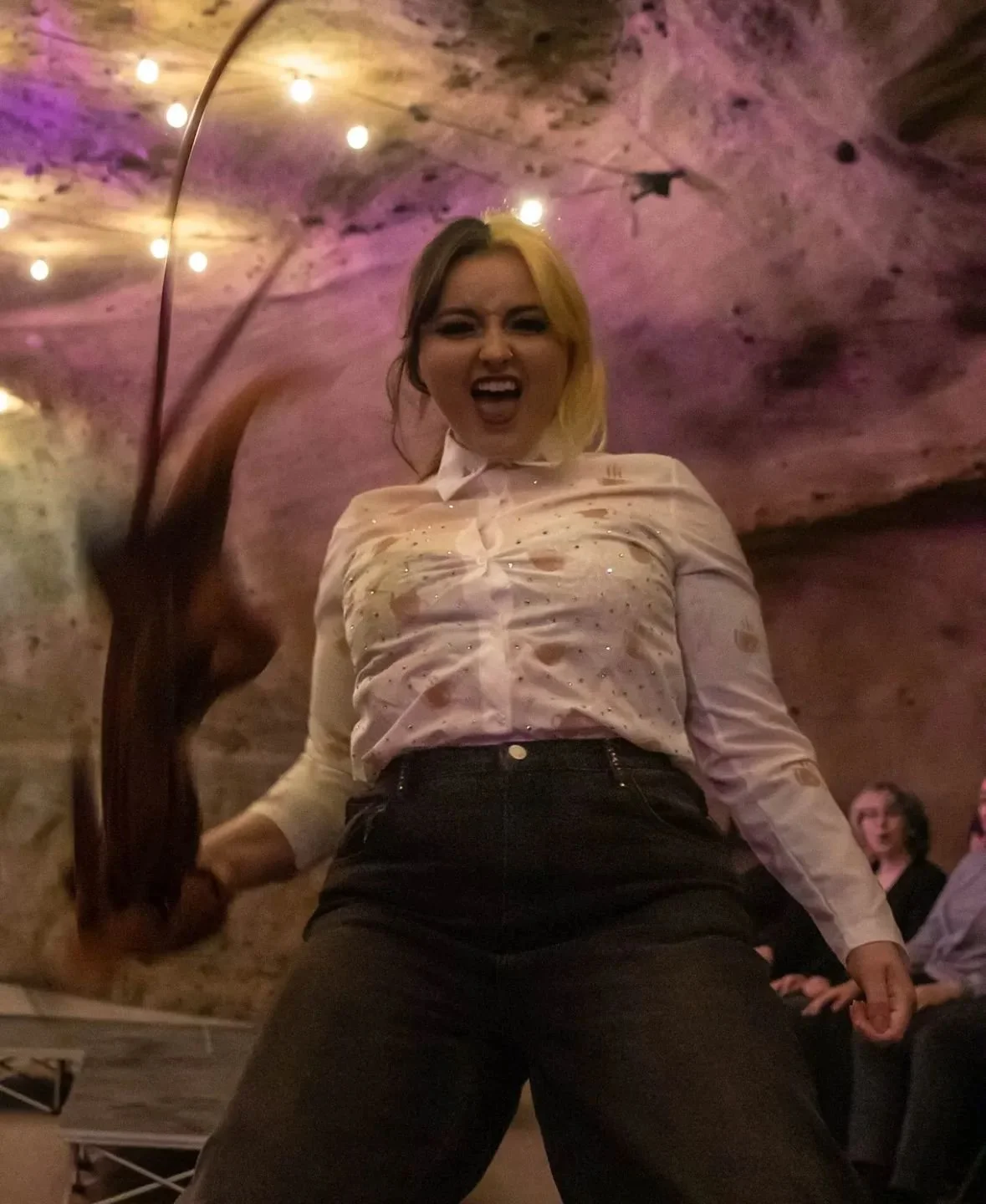 A woman wearing a light-colored, sparkly shirt and dark pants, appears to be in a lively setting, possibly a club or bar, with a colorful, rock-like ceiling and warm lighting. She has an expressive, energetic pose and facial expression.