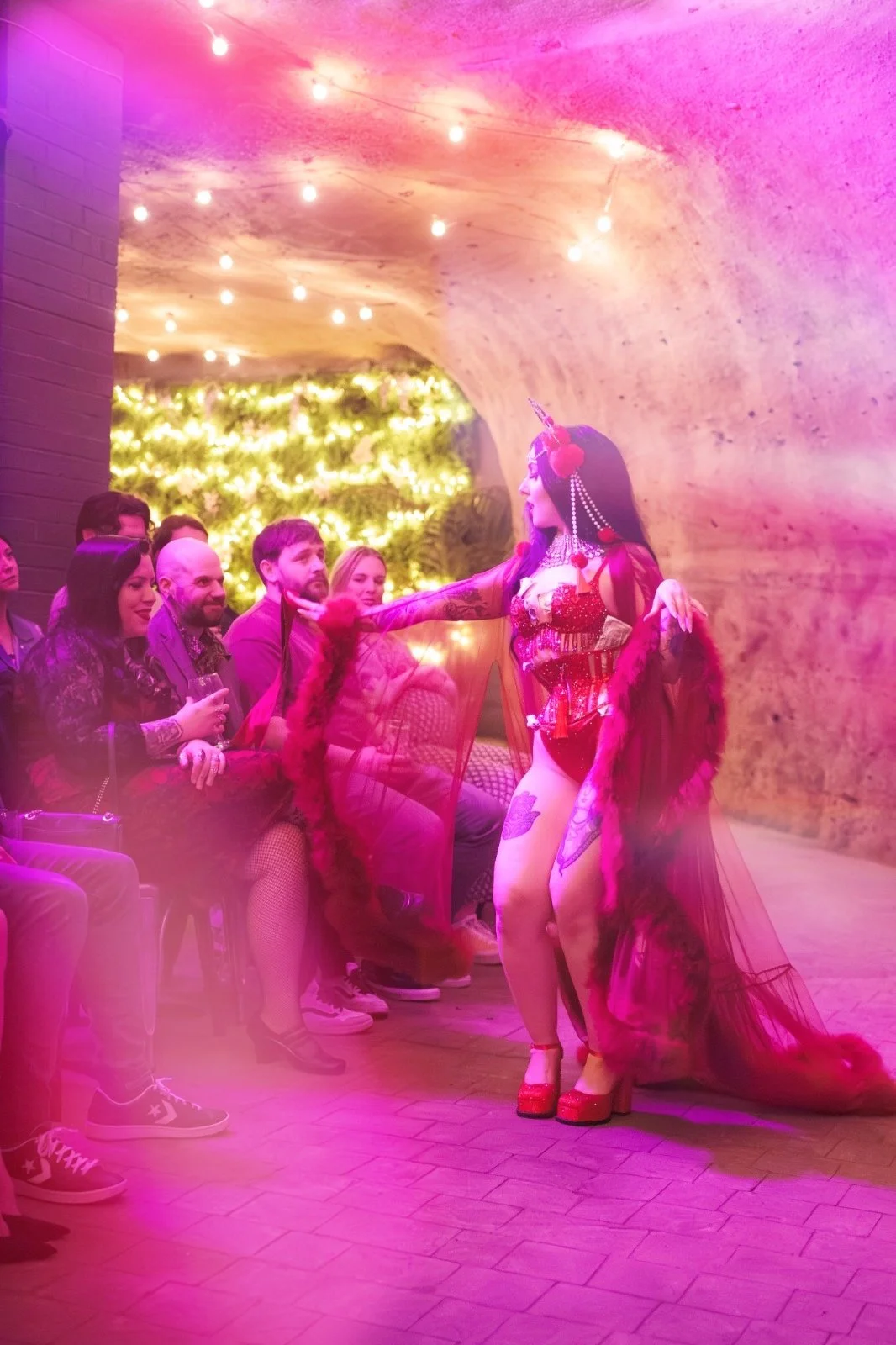 A performer dressed as a unicorn with a red and purple costume, long black hair, and colorful accessories interacts with an audience in a dimly lit room with pink and purple lighting, string lights, and a decorated wall.