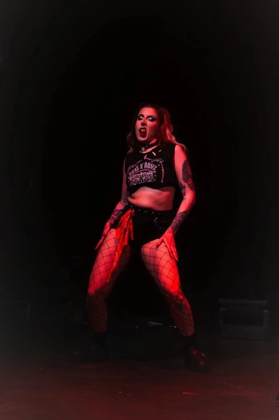 A woman performing on stage with a black background, wearing a ripped sleeveless shirt, fishnet stockings, and black shoes, with tattoos on her arms, and makeup with dark lipstick, striking a fierce pose.
