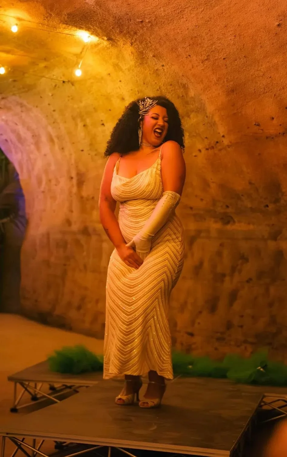 A woman in a gold dress and gloves is standing on a small stage, laughing and posing with one hand clutching her dress. She has curly hair and is wearing a decorative headpiece, in a warmly lit underground tunnel or cellar setting.