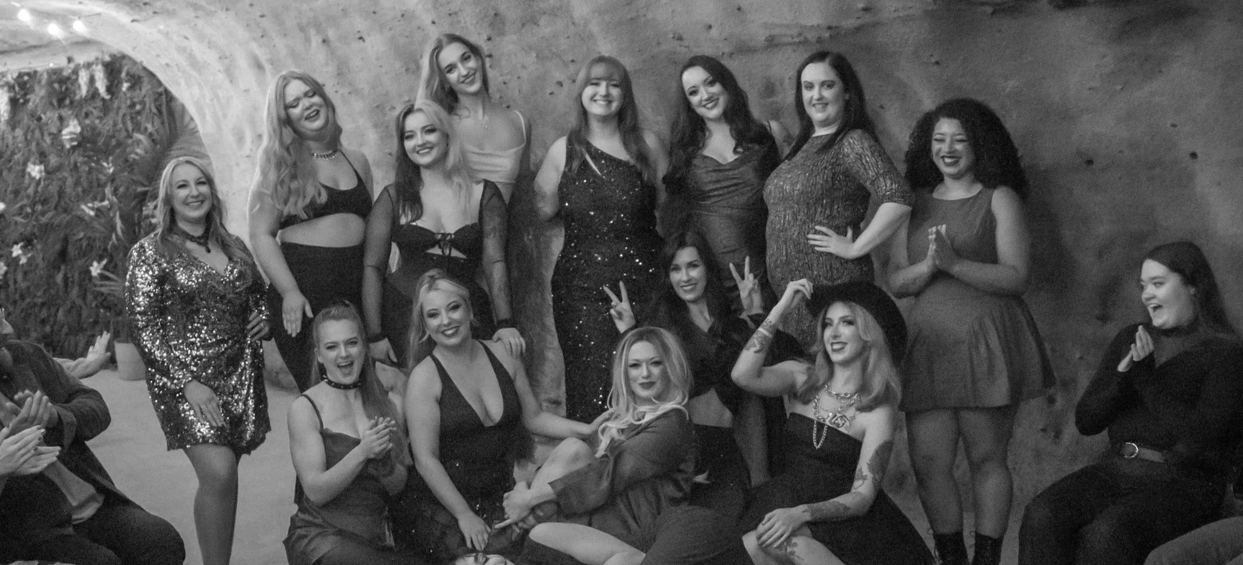A group of mostly women in glamorous attire, smiling and posing together in front of a textured wall at a social gathering or party.