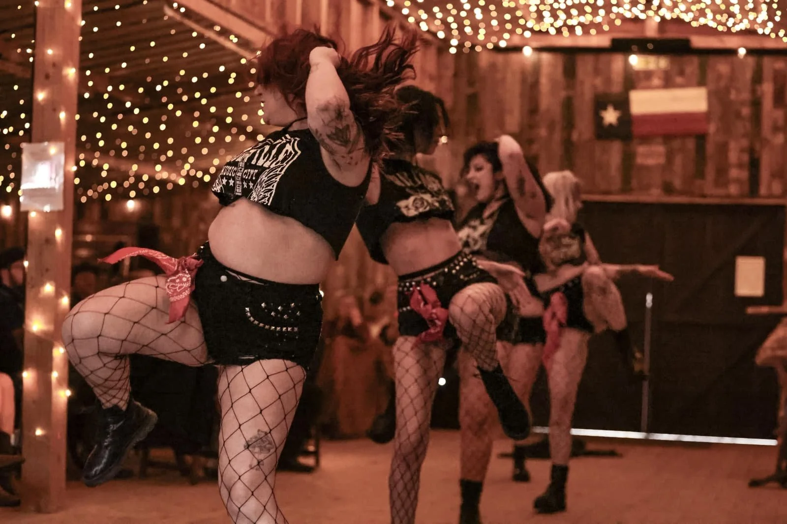Women in fishnet stockings and short shorts dancing with high knees in a rustic, dimly lit venue decorated with string lights and a vintage American flag on the wall.