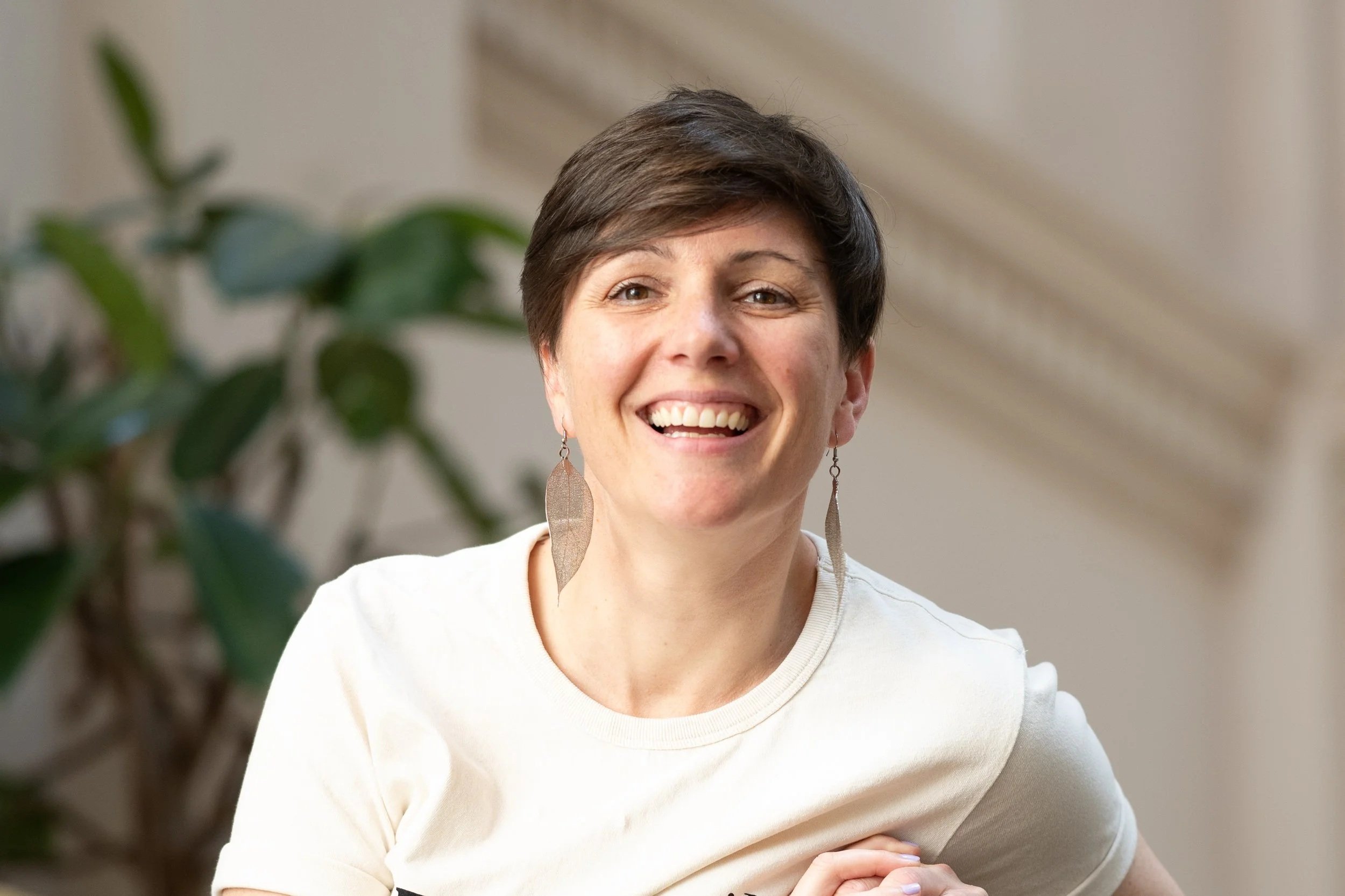A smiling woman with short brown hair and fun earrings, wearing a white top, sitting indoors near a plant with green leaves.