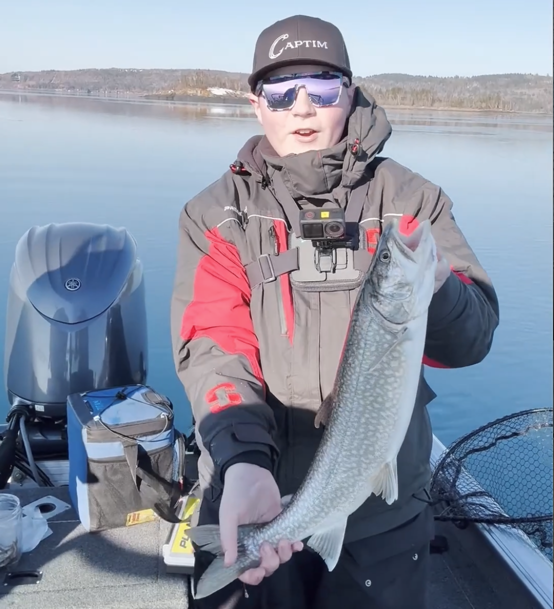 Person in outdoor fishing gear holding a large fish on a boat on a calm lake with hills in the background.