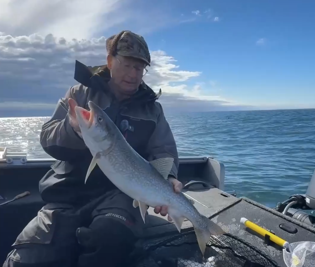 Man in outdoor gear holding a large fish on a boat in open water, with a cloudy sky and horizon in the background.