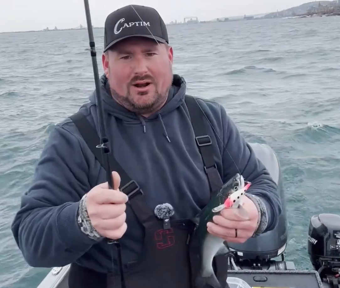 A man on a boat holding a fish and a fishing lure while giving a thumbs-up. He is wearing a black cap and hoodie, with an ocean and city skyline in the background.