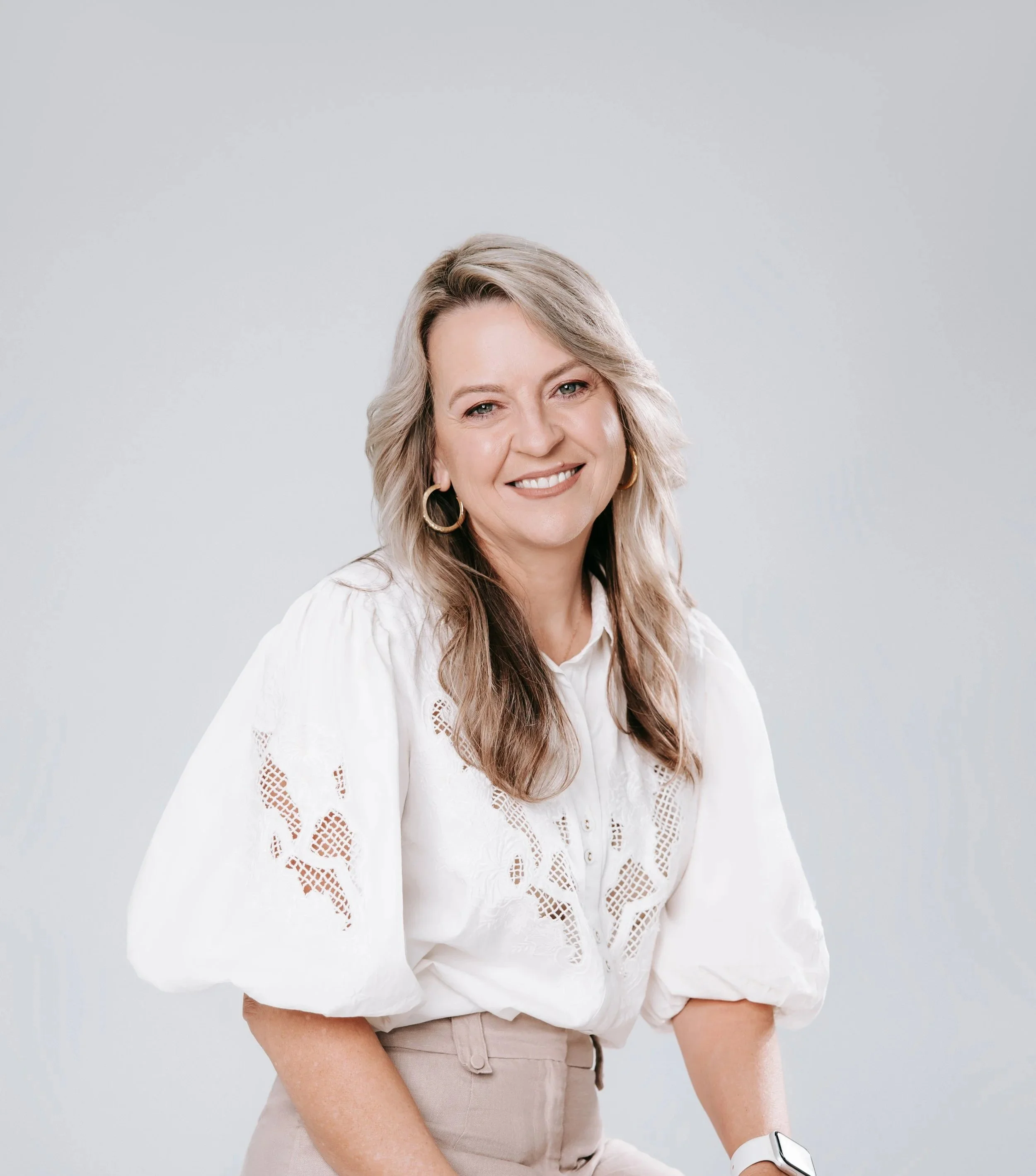 A Picture featuring Psychologist Kerstin Anderson Ridge with blonde wavy hair, wearing large hoop earrings, a white lace blouse with eyelet details, and beige pants, against a plain gray background.