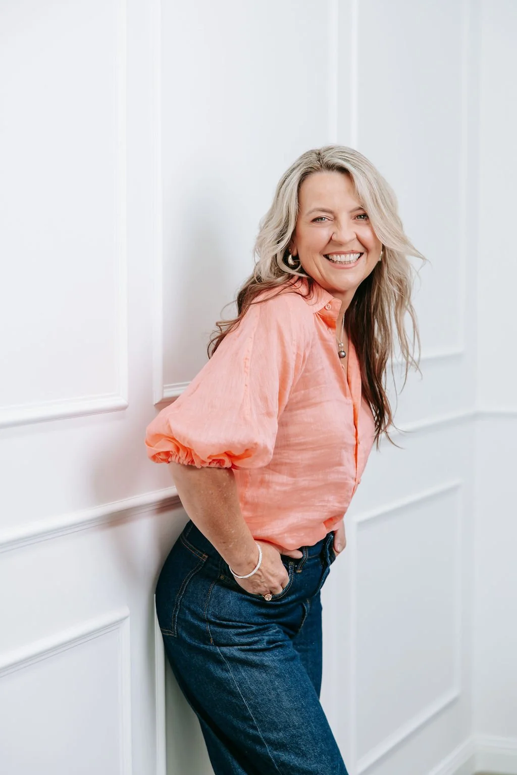 A Picture featuring Psychologist Kerstin Anderson Ridge with blonde hair in loose waves, wearing a peach-colored blouse with puffed sleeves and blue jeans, is smiling and leaning against a white paneled wall.